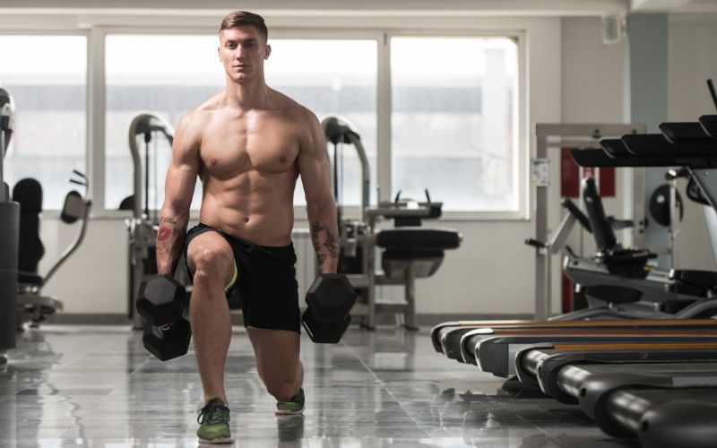 Shirtless man doing lunges holding dumbbells in each hand.