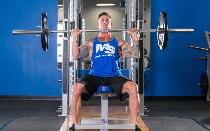 Muscular male athlete wearing blue tank doing a seated barbell shoulder press in the gym