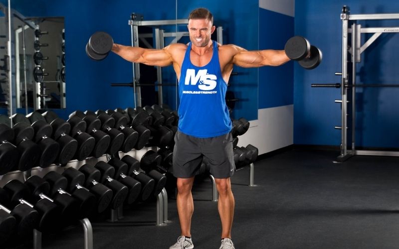 Muscular male athlete wearing blue tank doing lateral raises in the gym